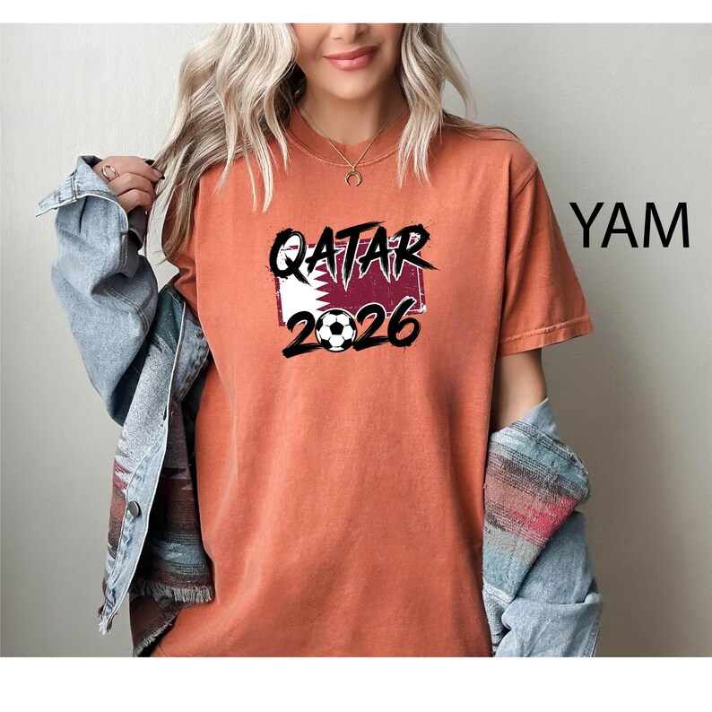 May include: An orange t-shirt with the text "QATAR 2026" in black, with a soccer ball graphic. The shirt features a design incorporating the Qatar flag. The shirt is a short-sleeved, casual style.