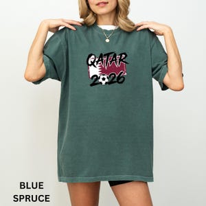 May include: Blue spruce t-shirt with the text "QATAR 2026" in black and white, along with a soccer ball graphic. The shirt features the Qatar flag in the design.