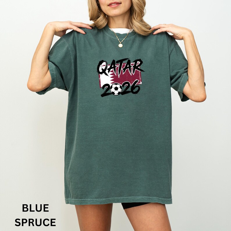 May include: Blue spruce t-shirt with the text "QATAR 2026" in black and white, along with a soccer ball graphic. The shirt features the Qatar flag in the design.
