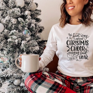 May include: A white long-sleeved shirt with black text that reads "The best way to spread Christmas cheer is singing loud for all to hear". The person wearing the shirt is holding a white mug and sitting in front of a snowy Christmas tree.