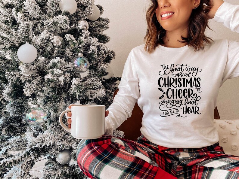 May include: A white long-sleeved shirt with black text that reads "The best way to spread Christmas cheer is singing loud for all to hear". The person wearing the shirt is holding a white mug and sitting in front of a snowy Christmas tree.