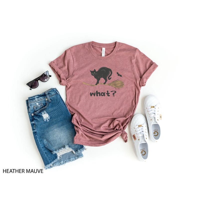 May include: Heather mauve t-shirt with a black cat on a broom and the word "what?" printed on the front. The shirt is styled with denim shorts, sunglasses, and white sneakers. A casual outfit for Halloween.