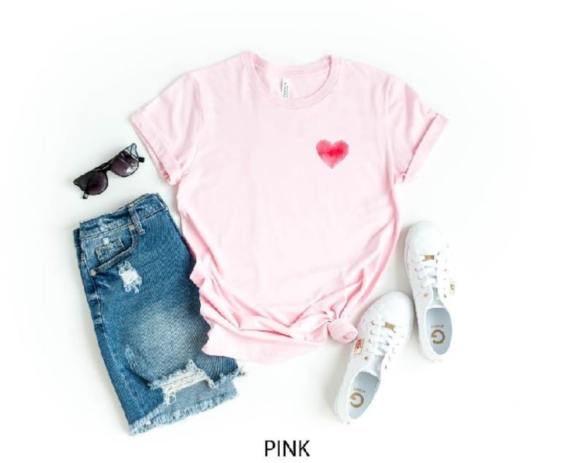May include: A pink t-shirt with a red watercolor heart design. The shirt is paired with distressed denim shorts and white sneakers.