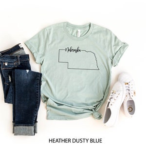 May include: A heather dusty blue t-shirt with a black outline of the state of Nebraska and the state's name in script. The shirt is paired with dark blue jeans and white sneakers. The text "HEATHER DUSTY BLUE" is at the bottom.