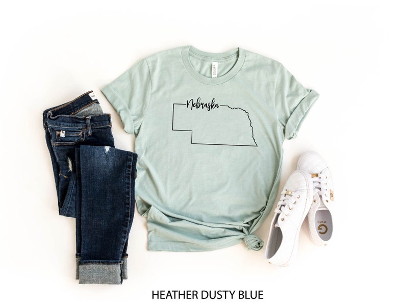 May include: A heather dusty blue t-shirt with a black outline of the state of Nebraska and the state's name in script. The shirt is paired with dark blue jeans and white sneakers. The text "HEATHER DUSTY BLUE" is at the bottom.