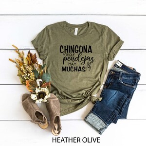 May include: A green t-shirt with the text "CHINGONA PORQUE HAY PENDEJAS MUCHAS" in black lettering. The shirt is on a white wooden surface with a pair of brown shoes, a bouquet of dried flowers, and a pair of blue jeans.