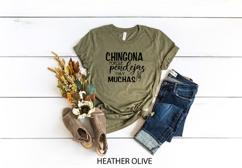 May include: A green t-shirt with the text "CHINGONA PORQUE HAY PENDEJAS MUCHAS" in black lettering. The shirt is on a white wooden surface with a pair of brown shoes, a bouquet of dried flowers, and a pair of blue jeans.