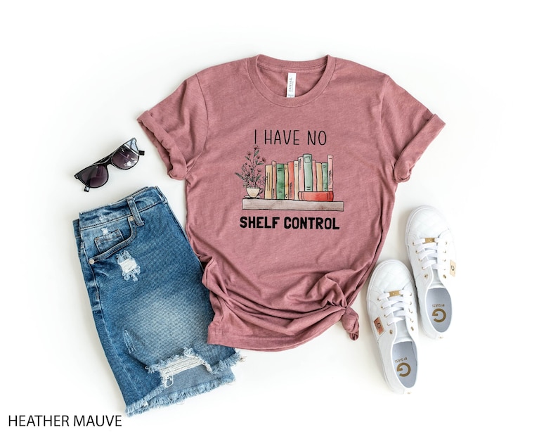 Pu&ograve; includere: Una t-shirt color malva con la scritta "I have no shelf control" e una grafica di una libreria con libri e una pianta. La t-shirt &egrave; mostrata su uno sfondo bianco con un paio di occhiali da sole, una gonna di jeans e scarpe da ginnastica bianche.
