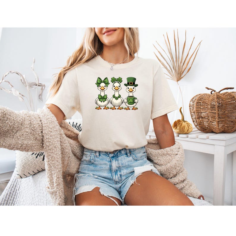May include: Cream-colored t-shirt featuring three cartoon geese, each adorned with St. Patrick's Day accessories. One goose wears a green bow, another a shamrock, and the third a green hat and holds a mug. The geese are centered on the shirt.