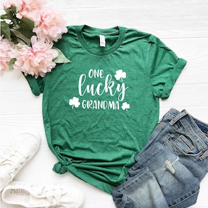 Puede incluir: Camiseta verde con texto blanco que dice "One lucky grandma" con dise&ntilde;os de tr&eacute;boles. La camiseta est&aacute; doblada y puesta sobre una superficie blanca con un par de zapatos blancos y unos vaqueros azules.
