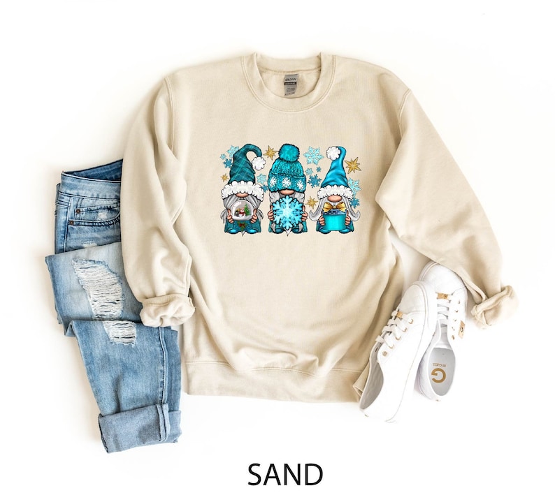 Op de afbeelding: Een beige sweatshirt met een afbeelding van drie gnomen die blauwe en witte wintermutsen dragen en een sneeuwvlok en een cadeaudoos vasthouden. De gnomen zijn omringd door sneeuwvlokken. Het sweatshirt is op een witte achtergrond.