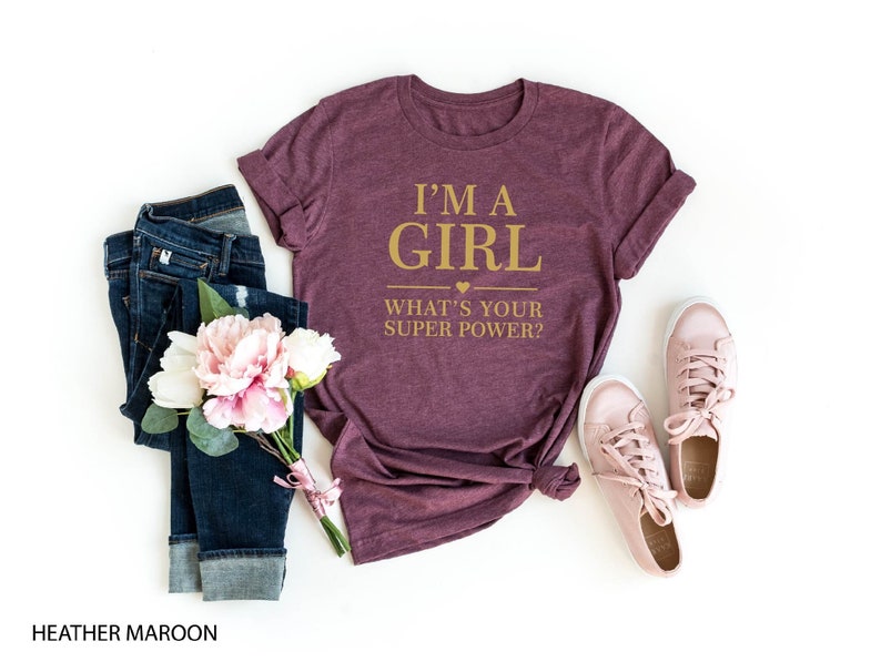 May include: A heather maroon t-shirt with the text "I'm a girl. What's your superpower?" in gold lettering.