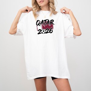 May include: White t-shirt with the text "QATAR 2026" in black and burgundy. The design includes a soccer ball and a burgundy rectangle with a pattern. The shirt is a casual, oversized fit.