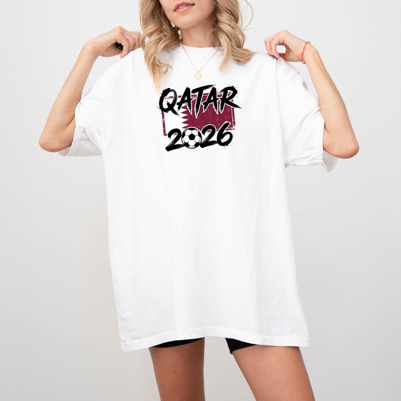 May include: White t-shirt with the text "QATAR 2026" in black and burgundy. The design includes a soccer ball and a burgundy rectangle with a pattern. The shirt is a casual, oversized fit.