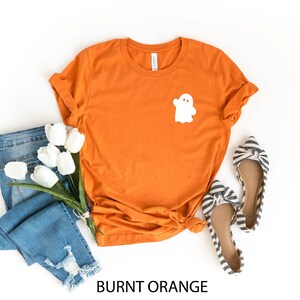 May include: Burnt orange t-shirt with a white ghost graphic. The shirt is paired with blue jeans and striped flats.