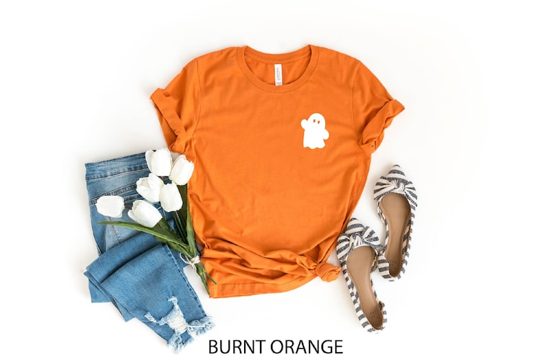 May include: Burnt orange t-shirt with a white ghost graphic. The shirt is paired with blue jeans and striped flats.