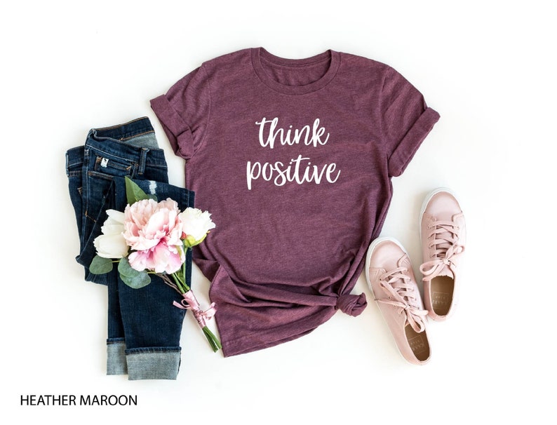 May include: Heather maroon t-shirt with the text "think positive" in white lettering. The shirt is shown with blue jeans, a bouquet of pink and white flowers, and pink trainers.
