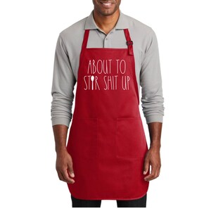 May include: Red kitchen apron with two pockets and white text that reads "About to Stir Shit Up" with a spoon graphic.