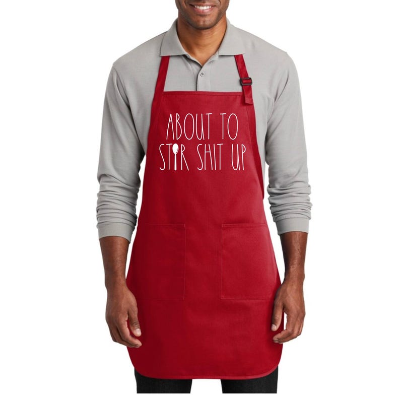 May include: Red kitchen apron with two pockets and white text that reads "About to Stir Shit Up" with a spoon graphic.