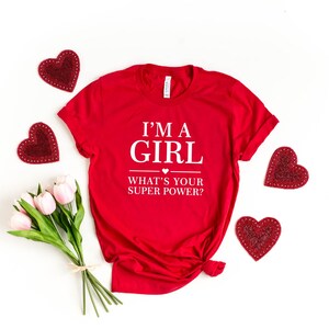 May include: Red t-shirt with white text that reads "I'm a girl what's your super power?" with a small heart symbol above the word "super". The shirt is surrounded by red heart-shaped decorations.