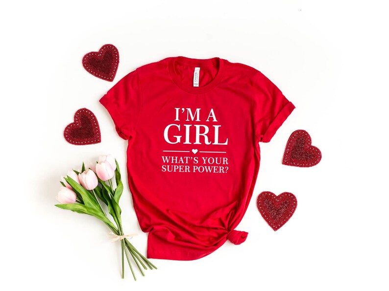 May include: Red t-shirt with white text that reads "I'm a girl what's your super power?" with a small heart symbol above the word "super". The shirt is surrounded by red heart-shaped decorations.