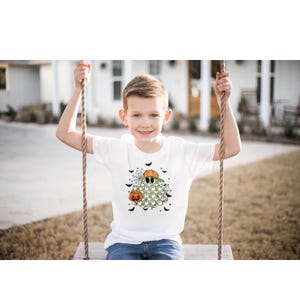 May include: White t-shirt featuring a Halloween ghost design. The ghost wears a baseball cap and sunglasses, holding a pumpkin. Black bats and spiderwebs surround the ghost. The shirt is worn by a child on a swing.