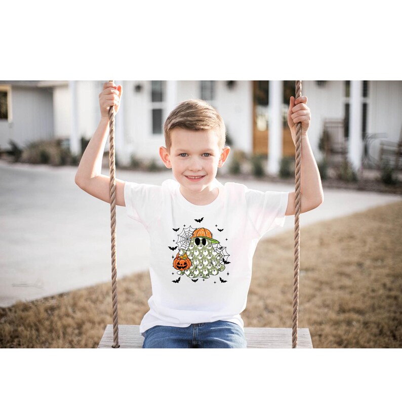 May include: White t-shirt featuring a Halloween ghost design. The ghost wears a baseball cap and sunglasses, holding a pumpkin. Black bats and spiderwebs surround the ghost. The shirt is worn by a child on a swing.