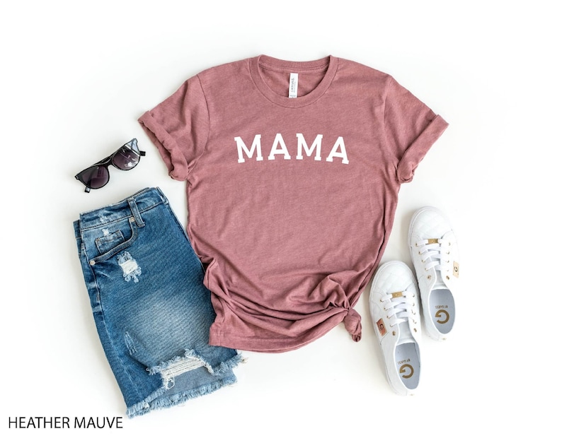 May include: A heather mauve t-shirt with the word "MAMA" printed in white on the front. The t-shirt is paired with denim shorts and white sneakers.
