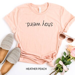 May include: A peach colored t-shirt with the text "Stay Weird." printed on the front. The shirt is laid flat on a white background with a pair of gray shoes, sunglasses, and a bouquet of pink roses.