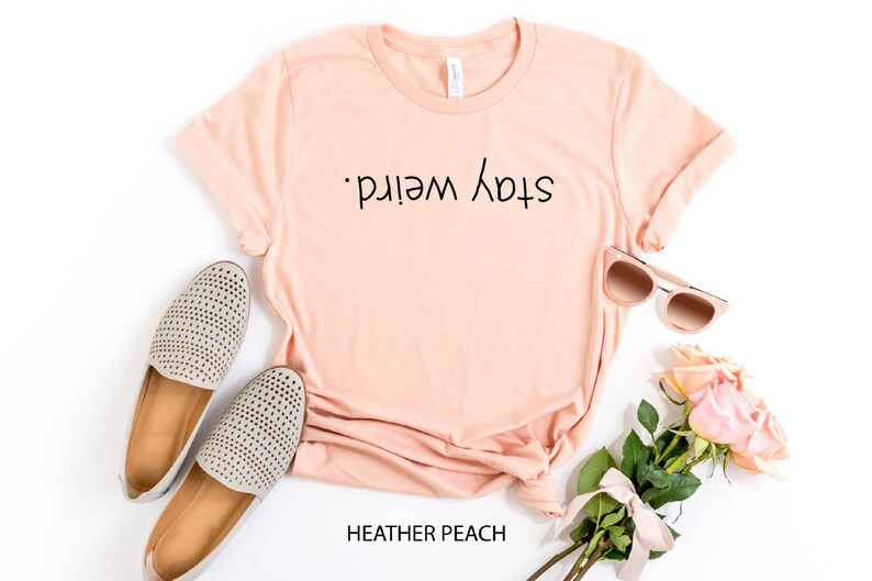 May include: A peach colored t-shirt with the text "Stay Weird." printed on the front. The shirt is laid flat on a white background with a pair of gray shoes, sunglasses, and a bouquet of pink roses.