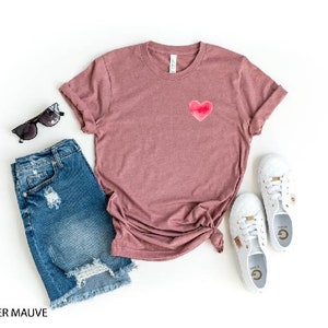 May include: A heather mauve t-shirt with a pink heart graphic. The t-shirt is paired with denim shorts and white sneakers.
