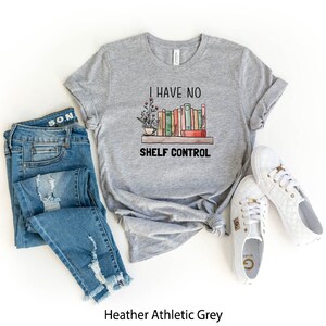 Pu&ograve; includere: Una t-shirt grigia atletica heather con il testo "I have no shelf control" e una grafica di una libreria con libri e piante. La t-shirt &egrave; abbinata a jeans blu e scarpe da ginnastica bianche.