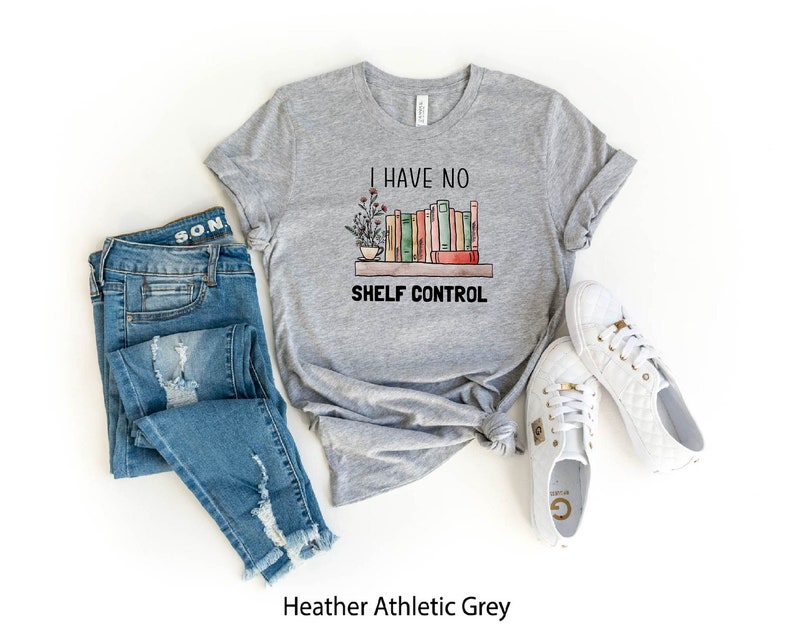 Pu&ograve; includere: Una t-shirt grigia atletica heather con il testo "I have no shelf control" e una grafica di una libreria con libri e piante. La t-shirt &egrave; abbinata a jeans blu e scarpe da ginnastica bianche.