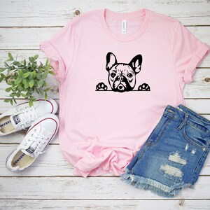 Puede incluir: Una camiseta rosa con una silueta negra de un bulldog franc&eacute;s asom&aacute;ndose por la parte superior. El bulldog tiene las patas en el borde de la camiseta. La camiseta est&aacute; colocada sobre una superficie de madera blanca con un par de zapatillas blancas y unos pantalones cortos de mezclilla azul.