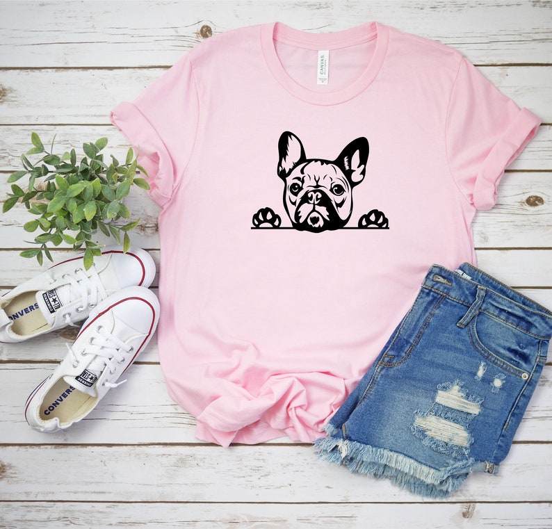 Puede incluir: Una camiseta rosa con una silueta negra de un bulldog franc&eacute;s asom&aacute;ndose por la parte superior. El bulldog tiene las patas en el borde de la camiseta. La camiseta est&aacute; colocada sobre una superficie de madera blanca con un par de zapatillas blancas y unos pantalones cortos de mezclilla azul.
