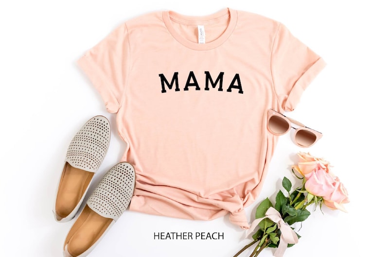 May include: A peach colored t-shirt with the word "MAMA" printed in black on the front. The shirt is laid flat on a white background with a pair of gray shoes, sunglasses, and a bouquet of pink roses.