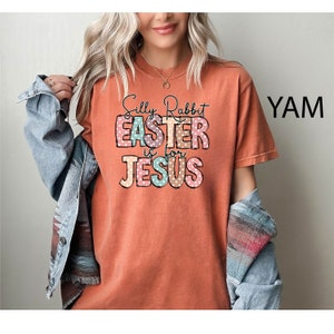 Może przedstawiać: Koszulka w kolorze rdzy z nadrukiem graficznym, na kt&oacute;rym widnieje napis "Silly Rabbit Easter is for Jesus" w kolorowych literach z białymi kropkami.