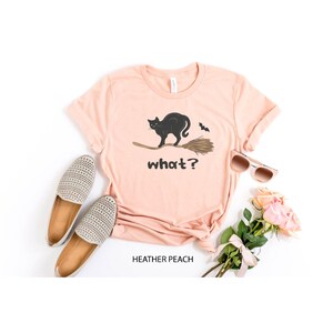 May include: A heather peach t-shirt featuring a black cat riding a broom with a bat and the word "what?" in black. The shirt is paired with beige shoes, sunglasses, and a bouquet of pink roses.