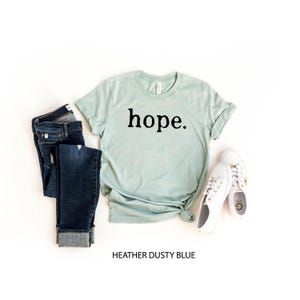 May include: A light blue t-shirt with the word "hope" printed in black on the front. The shirt is shown on a white background with a pair of blue jeans and white shoes.