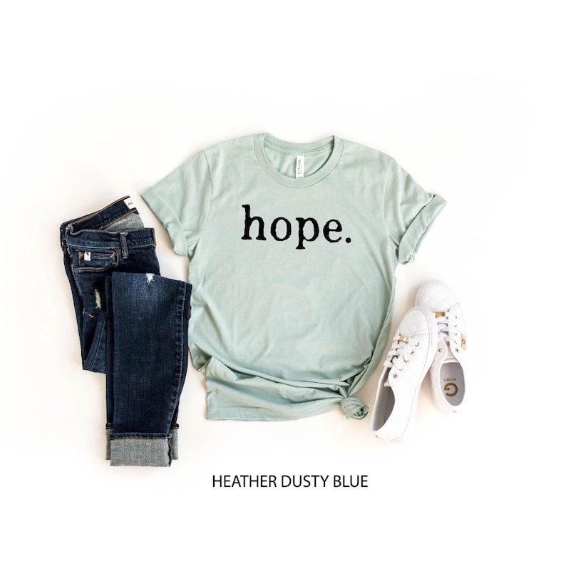 May include: A light blue t-shirt with the word "hope" printed in black on the front. The shirt is shown on a white background with a pair of blue jeans and white shoes.