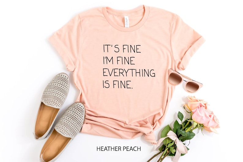 May include: A peach-colored t-shirt with the text "It's fine I'm fine Everything is fine." printed in black. The shirt is laid flat on a white surface with a pair of gray shoes, sunglasses, and pink roses.