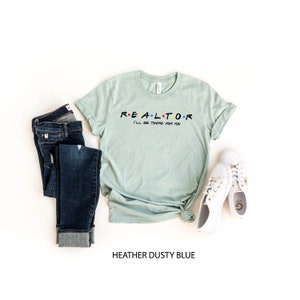 May include: A heather dusty blue t-shirt with the word "REALTOR" in colorful letters, and the phrase "I'll be there for you" below it. The shirt is paired with dark blue jeans and white sneakers.