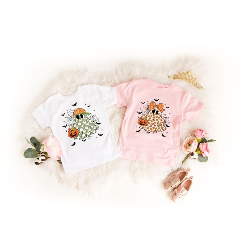 May include: Two short-sleeved Halloween-themed t-shirts. One is white with a ghost wearing sunglasses and a pumpkin. The other is pink with a ghost wearing a bow and holding a pumpkin. Both shirts have spiderwebs and bats.