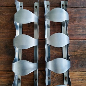 May include: Two brushed metal wine racks designed to hold multiple bottles. Each rack has a series of curved holders and mounting holes for wall installation. The racks are displayed against a dark wood background.