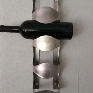 May include: A brushed metal wine rack mounted on a wall, holding a dark wine bottle. The rack has a vertical design with three curved cradles to secure the bottles. The rack is made of stainless steel.
