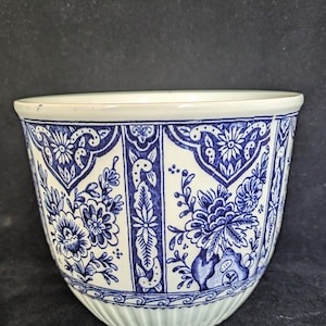 May include: A blue and white ceramic planter with a floral pattern. The planter has a repeating design of blue flowers and foliage on a white background. The rim and base have a simple, clean design.