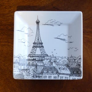 May include: A white square ceramic dish with a black and white line drawing of the Eiffel Tower and Parisian rooftops. The drawing includes clouds and birds in the sky.