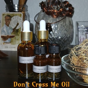 以下が含まれることがあります： 金色のキャップと白いスポイトが付いた「Don't Cross Me」オイルのガラス瓶が3本。瓶は濃い茶色の液体で満たされ、ハーブと花が入っています。瓶のラベルには「Don't Cross Me Soular Space」と書いてあります。