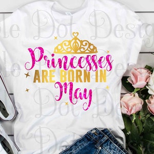 May include: White t-shirt with the text "Princesses are born in May" in pink and gold glitter lettering with a gold crown above the text. The shirt is lying on a white wooden surface with a pair of white trainers and a bouquet of pink roses.