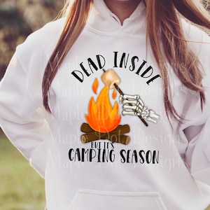 May include: White hoodie with a graphic of a skeleton roasting a marshmallow over a campfire. The text reads "Dead inside but it's camping season".